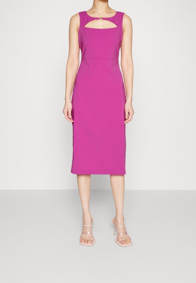 Femme portant une robe magenta sans manches, longueur genou, avec une découpe au niveau de la poitrine et des sandales à talons hauts à brides transparentes.