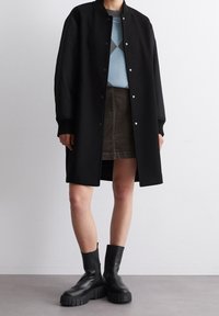 Cappotto lungo nero con collo arrotondato, bottoni a scatto, polsini a coste, indossato sopra un maglione blu e una gonna in velluto a coste marrone. Stivali platform neri.