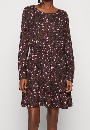 Vrouw draagt een bruine jurk met lange mouwen met roze en zwarte luipaardprint en een strakke elastische taille, staande voor een effen achtergrond.