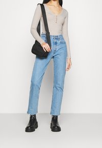 Nainen pukeutuneena beigeen ribbineulottuun pitkähihapaidankaltaiseen paitaan, vaalean sinisiin korkeavyötäröisiin farkkuihin, joiden helmat ovat rispaantuneet, mustiin saappaisiin ja mustaan olkalaukkuun.