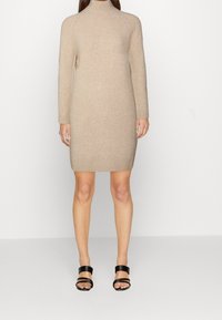 Abito lavorato a maglia beige con collo alto e maniche lunghe. Dettaglio sul bordo a coste. Abbinato a sandali neri con tacco e dettagli a fascetta.
