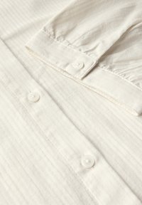 Bílá košile na knoflíky vyrobená z texturované, žebrované látky. Má límeček s špičkami a dva viditelné knoflíky na přední léze.