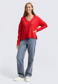 Pull rouge en tricot avec un col en V, coupe décontractée et poignets côtelés, associé à un jean bleu clair à jambes droites et des baskets blanches.