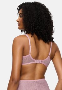 Ženska s kodrastimi lasmi nosi svetlo rožnato usklajeno kompleto modrčka in hlačic z visokim pasom, izdelano iz subtilno vzorčaste tkanine, prikazana zadaj.