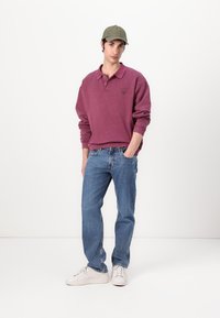 Sweatshirt bordeaux avec un col et un logo sur la poitrine gauche, associé à un jean droit bleu et des sneakers blanches. Casquette vert olive.
