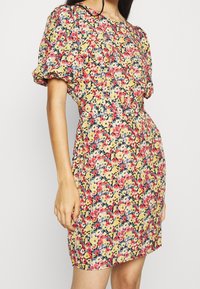 Blumenkleid mit einer Mischung aus rosa, gelben, blauen und grünen Blumen auf gelbem Hintergrund. Kurze Puffärmel, taillierte Silhouette und über knielange Länge.