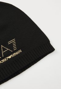 Zwarte gebreide beanie met een geribbelde omslag, versierd met een gouden logo en gestuikte letters, die een gestructureerd en stijlvol ontwerp biedt.