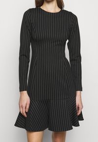 Černé šaty s vertikálními bílými pruhy, dlouhými rukávy, přiléhavým živůtkem a rozšířenou sukní. Materiál se zdá být hladký; má kulatý výstřih.