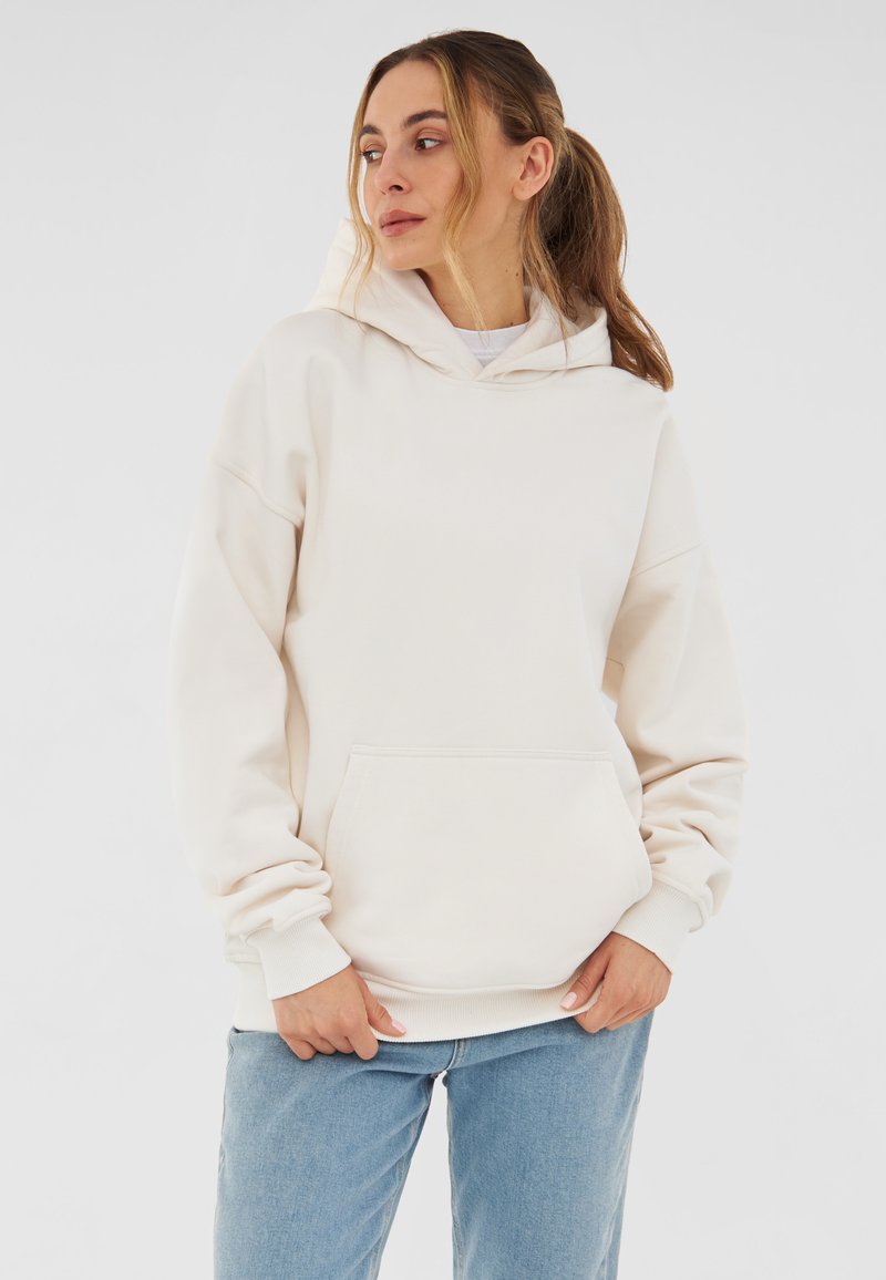 Sudadera color crema con un ajuste relajado, gran bolsillo frontal y capucha con cordón. Textura suave, puños y dobladillo acanalados. Combinada con vaqueros de color azul claro.