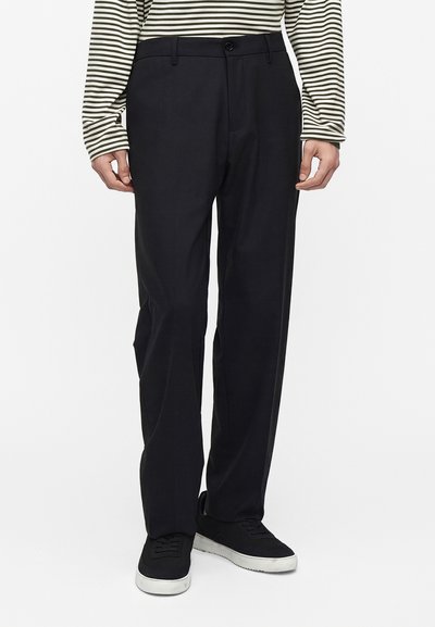 Personne portant un pantalon noir droit, des baskets noires avec des semelles blanches, et une chemise blanche à manches longues avec des rayures horizontales noires.