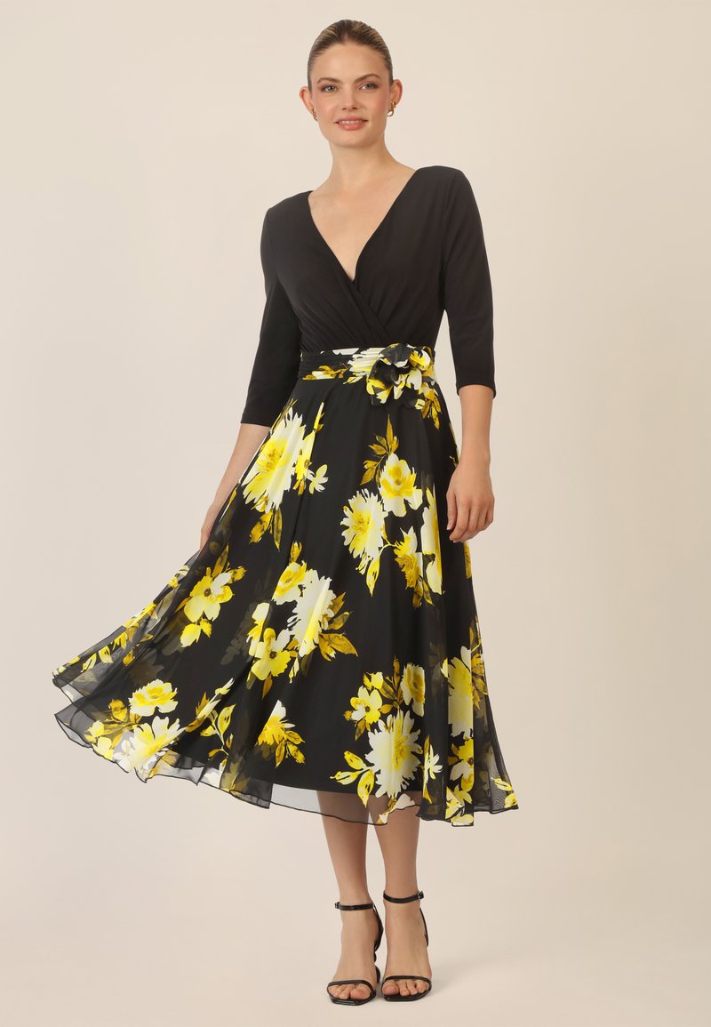 Zwarte bloemenjurk met een diepe V-hals en driekwartmouwen. Bevat gele en witte bloemen, een aansluitende taille en een vloeiende rok.