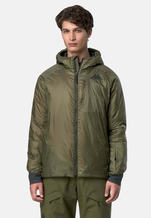 Giovane uomo che indossa una giacca a vento con cappuccio verde oliva e pantaloni cargo abbinati, in piedi contro uno sfondo chiaro uniforme.