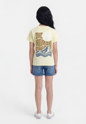 Girl with long black hair wearing a yellow The North Face t-shirt with mountain and sun graphic, denim shorts, and white sneakers, facing away.