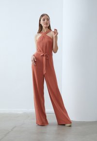 Terrakotta-farget jumpsuit med vide ben, halterneck og knyttet midje. Laget av strukturert stoff med en avslappet passform.