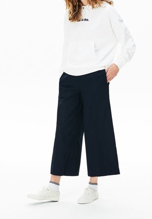 Persoon draagt een witte hoodie met de tekst "Lacoste", zwarte wijde cropped broek, witte sokken en witte sneakers, staand met één hand in de zak.