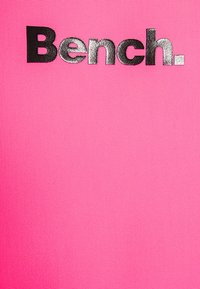Svetlo roza tekstil z gladko teksturo, ki na vrhu prikazuje izrazit črn natisnjen logotip "Bench." Preprost, drzno oblikovan.