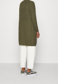 Oliivivihreä ribattu neuletakki, pitkä ja hieman pidempi takaa, yhdistettynä valkoisiin leveisiin housuihin ja oliivinvihreisiin Vans-kengissä.