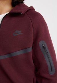 Burgundi kapucnis pulóver cipzáras zsebbel, szürke Nike logóval és fényes vízszintes csíkkal a mellkason. Sima tapintás, kapucnis kialakítás.