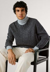 Maglione grigio a coste con colletto alto grigio chiaro e polsini a costine, indossato con pantaloni color crema, seduto su una sedia nera.