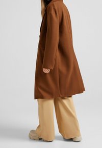 Brązowy długi płaszcz z mieszanki wełny z kołnierzem z klapami, pojedynczym zapięciem na guzik i kieszeniami po bokach; zestawiony z beżowymi szerokimi spodniami i masywnymi butami.