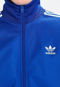 Blauwe ritsjack van gladde stof. Heeft een hoge kraag, witte strepen op de schouders en een geborduurd wit Adidas-logo op de borst.