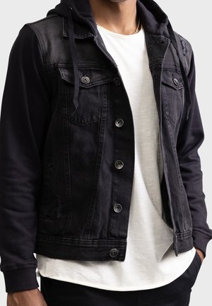 Homme portant une veste en jean noire avec manches de sweat à capuche par-dessus un t-shirt blanc, les mains partiellement dans les poches, sur un fond uni.