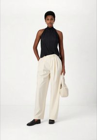 Woman standing in black sleeveless pleated top, high-neck, cream wide-leg trousers, black loafers, holding beige handbag with one hand.
