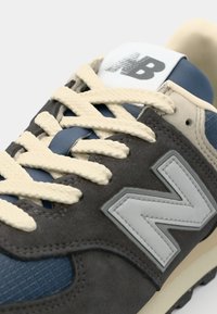 Zapatilla de ante gris con acentos de malla azul marino, cordones crema y un logotipo blanco en la lengüeta. Presenta una suela de goma texturizada y un diseño en capas.