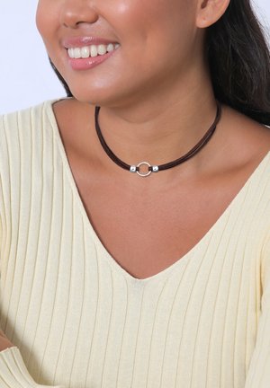 Brown suede choker with a circular silver metal accent. Smooth texture, layered design, worn with a light ribbed yellow top.