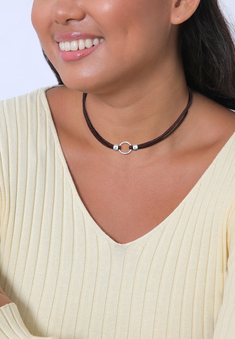 Choker en suède marron avec un accent circulaire en métal argenté. Texture lisse, design superposé, porté avec un haut jaune à côtes légères.