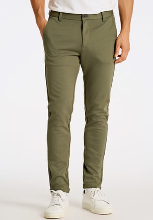 Chinos verdes de corte slim, fabricados en tela texturizada. Cuentan con cierre de botón, bolsillos laterales y dobladillos con puños. Combinados con zapatillas blancas.