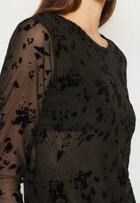 Svart genomskinlig långärmad topp med texturerat sammetmönster som har en subtil glans. Rund halsringning och figurnära design.