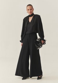 Čierne nohavice s širokými nohavicami v kombinácii s čiernou blúzkou s záhybmi. Doplnky zahŕňajú čiernu listovú kabelku a špicaté lodičky. Celý outfit má elegantnú saténovú textúru.