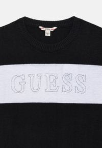 Juodas megztinis su baltu horizontalios juostos akcentu. Ant juostos iškeliama "GUESS" logotipas iš kristalų. Tekstūrinis banguotas apykaklės kraštas.