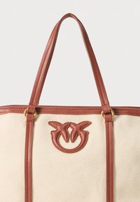 Tote bag din pânză cu curele din piele maro și un logo care prezintă două forme asemănătoare frunzelor. Culoare crem cu detalii subtile de textură.