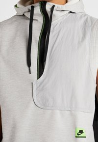 Hoodie sans manches gris clair avec une fermeture éclair noire, des accents verts et une poche appliquée. Le tissu est doux avec une texture subtile.