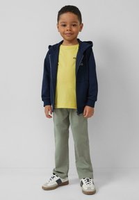 Marineblauwe zip-up hoodie over een gele t-shirt, gecombineerd met lichtgroene broek en witte sneakers. Kenmerkt zich door zachte stoffen en een casual ontwerp.