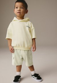 Hoodie jaune clair avec un graphisme, associé à un short cargo vert clair, et des baskets noires avec des semelles blanches et des bandes Velcro.