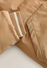  Tela de chaqueta color beige con una textura suave. El puño presenta una banda de punto con rayas en beige claro y blanco, complementada por un bolsillo lateral.