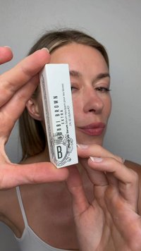 Donna che tiene una piccola scatola bianca di Bobbi Brown Extra lip serum vicino al viso con sfondo neutro.