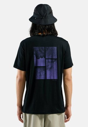 Person trägt schwarzen Bucket Hat und schwarzes T-Shirt mit abstraktem lila rechteckigem Design auf dem Rücken, steht vor einfachem Hintergrund.