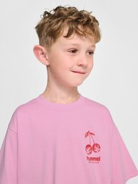 Młody chłopiec o kręconych blond włosach ubrany w różowy T-shirt z czerwonym nadrukiem wiśni i logo "hummel SPORTSWEAR" na piersi.