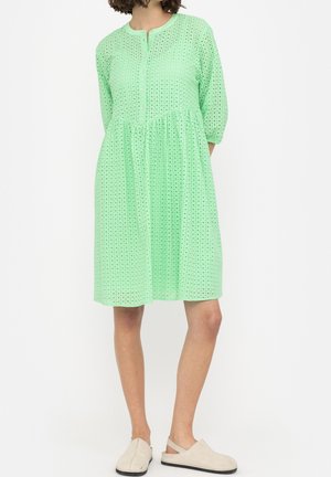 Frau trägt ein mintgrünes, knielanges Kleid mit Dreiviertelärmeln und einem quadratischen Lochmuster, kombiniert mit beigen Slippern.