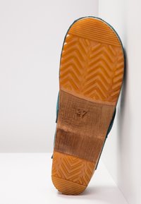 Semelle d'une chaussure bleu sarcelle avec une bande de roulement en caoutchouc orange et une section médiane en bois, taille 37 embossée, positionnée à la verticale contre un mur d'angle blanc.
