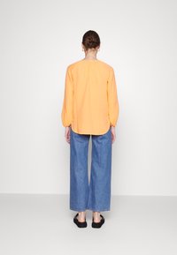 Blusa de manga longa laranja brilhante com detalhe de costas franzido, combinada com jeans de ganga azul de pernas largas e sapatos pretos de slip-on.