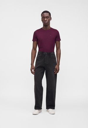 Homme debout portant un t-shirt ajusté violet foncé, un jean noir ample et des baskets blanches sur un fond blanc uni.