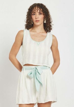 Ženska s kodrastimi lasmi, oblečena v belo brezrokavnik kratki top in ujemajoče se kratke hlače z visokim pasom ter svetlo modrim pasom, ki je zavezana v vozel.