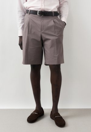 Mężczyzna w mauve plisowanych krótkich spodenkach do kolan, jasnoróżowej koszuli zapinanej na guziki włożonej do środka, czarnym pasku, brązowych mokasynach i srebrnej bransoletce.