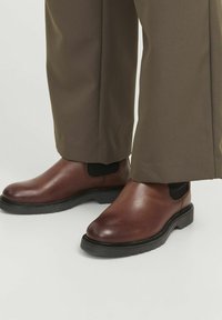 Bottes de cheville en cuir marron avec des panneaux élastiques sur les côtés, bout arrondi et semelle en caoutchouc, associées à un pantalon marron long et ample.