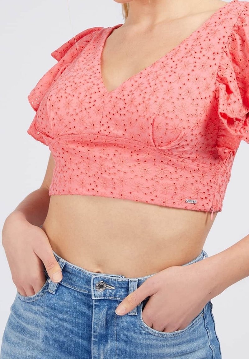 Femme portant un crop top rose texturé œillets avec des manches volantes et un jean bleu taille haute, mains partiellement dans les poches.